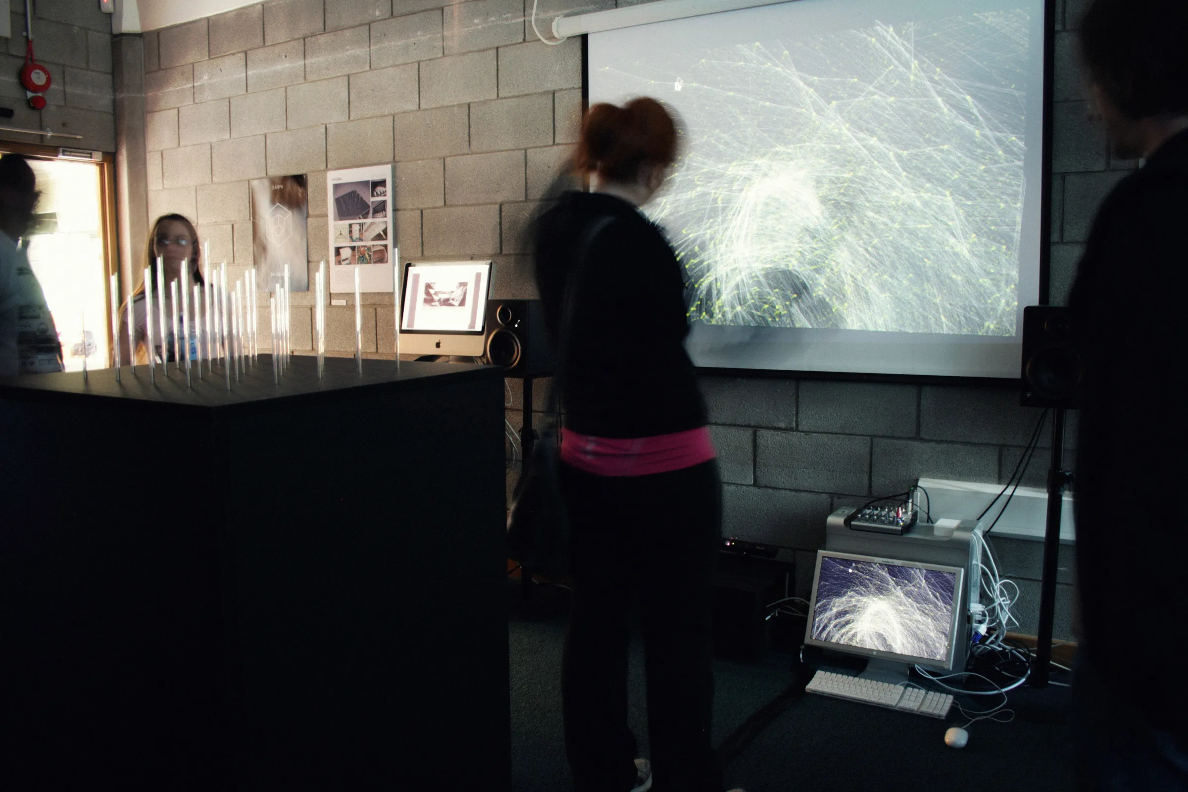 Visitors in a darkened room around the installation, with the pin plinth on the left and a projection of flowing lines on the wall