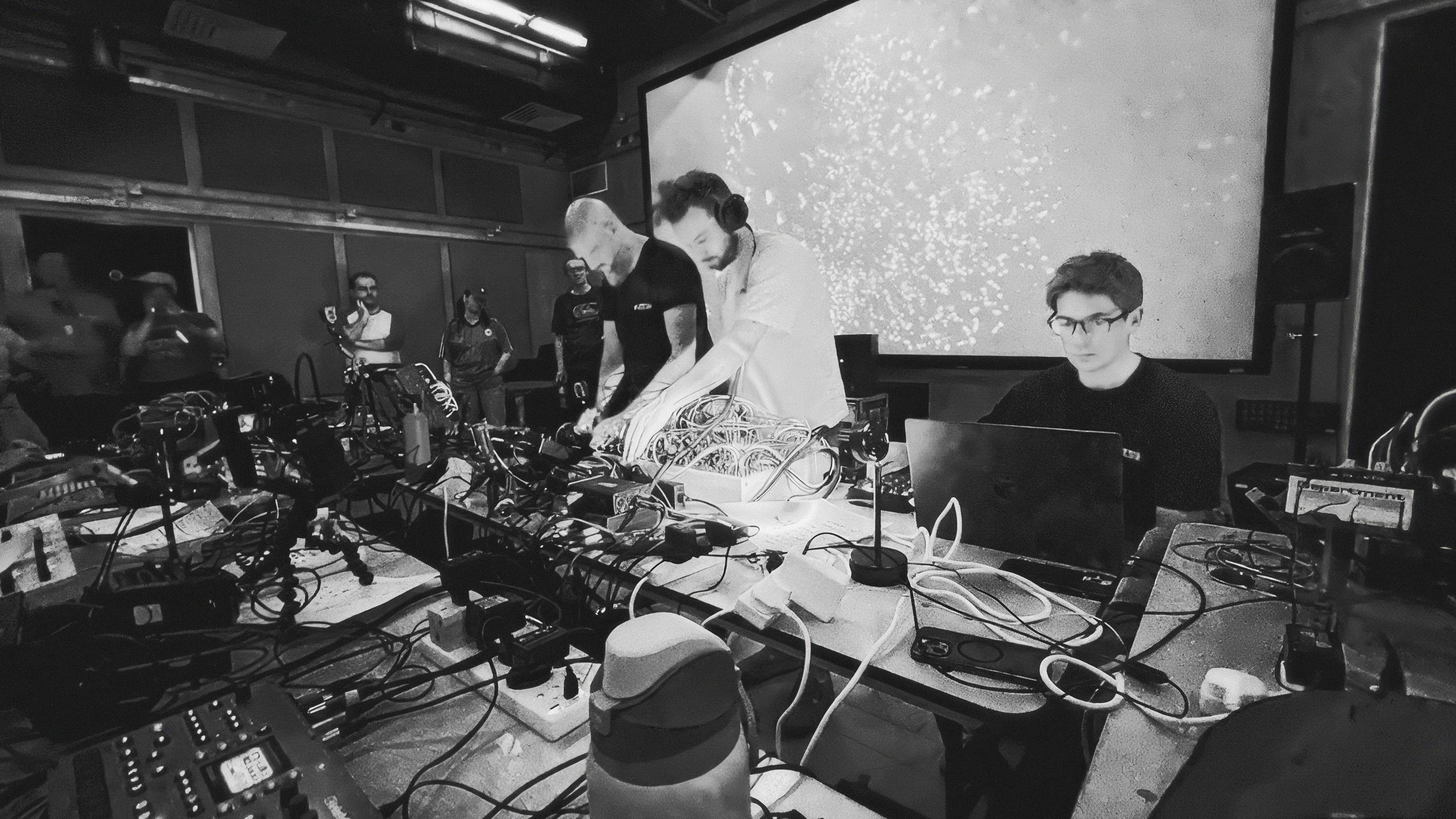 A black-and-white photo of two musicians patching a modular synth at a long table covered in cables and hardware, beside a person working on a laptop, in front of a large screen showing a shimmering particle field while an audience watches.