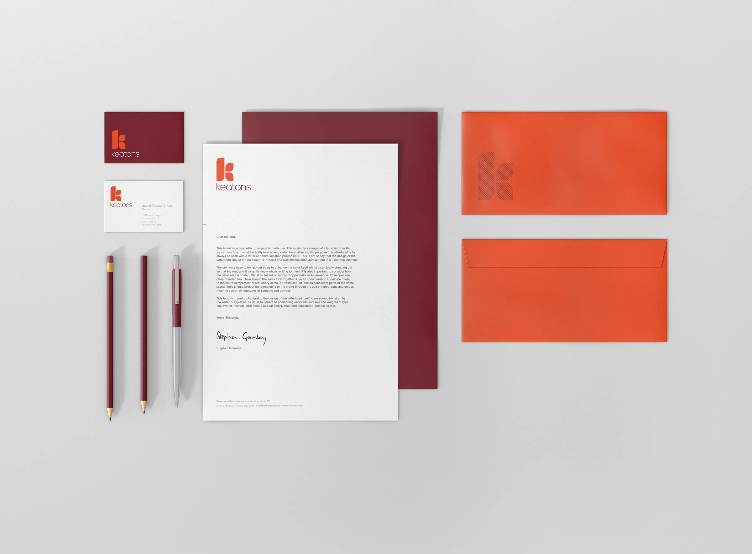 A flat lay of Keatons stationery, including burgundy and white business cards, a letterhead, a burgundy folder, burgundy pencils, and orange envelopes