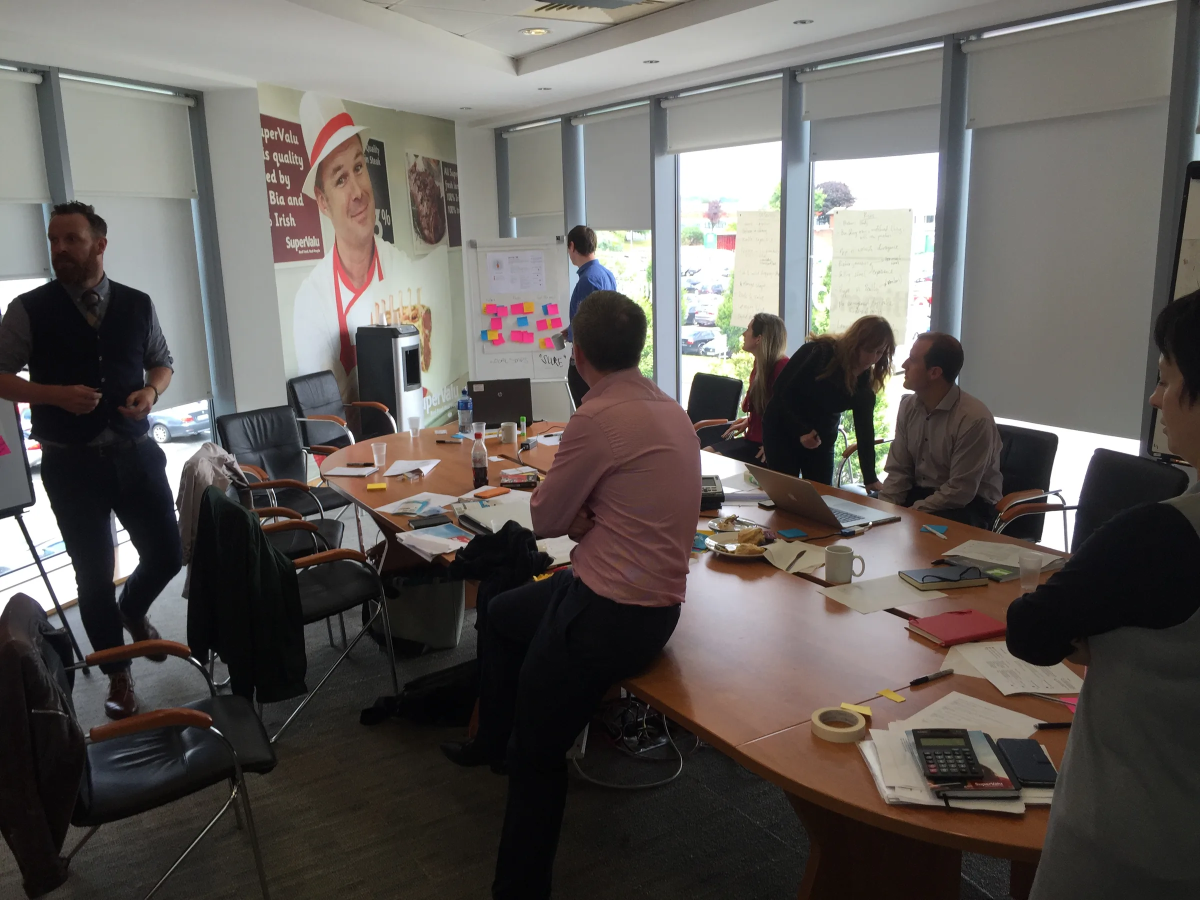 SuperValu team members around a boardroom table during a workshop, with sticky notes on the wall behind them